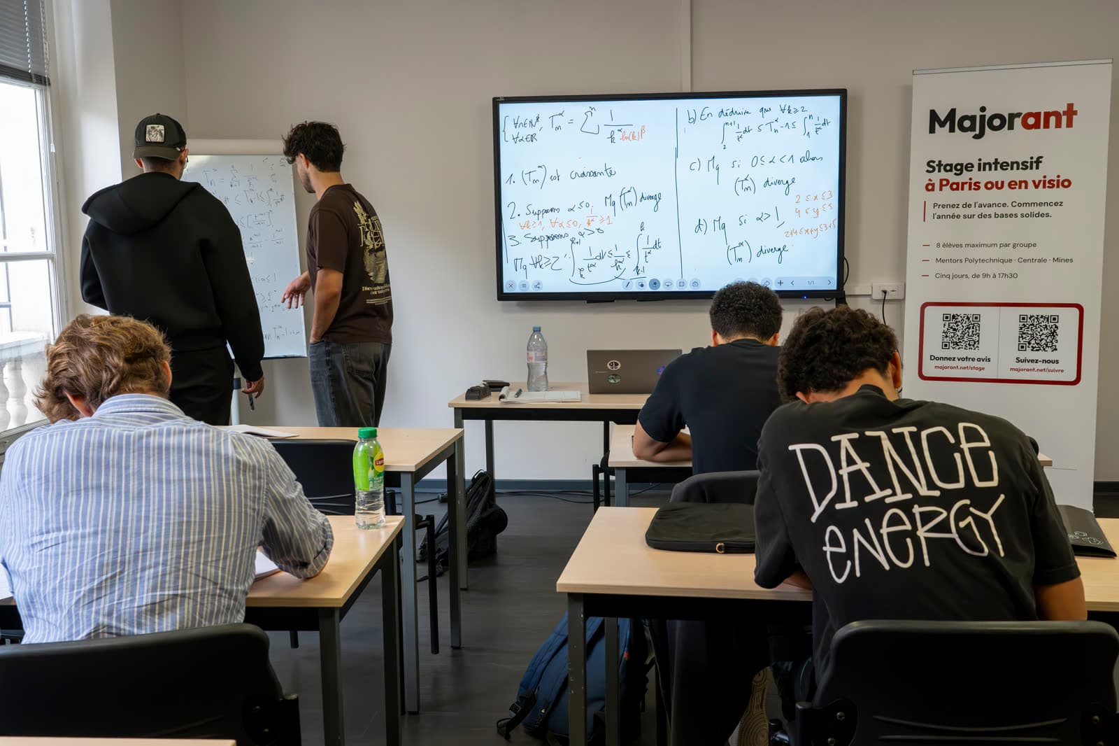 Une salle de cours avec des tables de travail et un tableau couvert d'exercices.